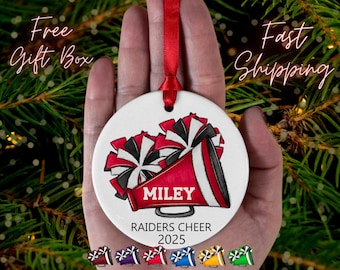 May include: A white ceramic ornament with a red ribbon for hanging. The ornament features a red and black cheerleader megaphone and pom-poms design. The name "MILEY" is printed on the megaphone, with "RAIDERS CHEER 2025" below.