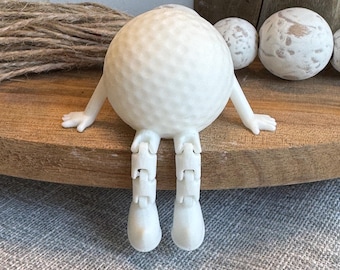 May include: White golf ball figurine with articulated legs and arms. The golf ball is sitting on a wooden surface.