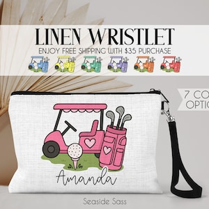 May include: White linen wristlet with a black zipper and wrist strap. The design features a pink golf cart, golf bag, and golf ball. The name "Amanda" is printed below the golf scene. The text "LINEN WRISTLET" and "7 COLOR OPTIONS" are also visible.