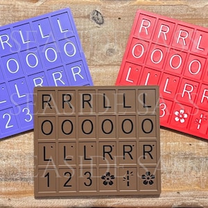 Może przedstawiać: Trzy kolorowe uchwyty na kafelki Scrabble w kolorze fioletowym, czerwonym i brązowym. Każdy uchwyt ma siatkę kwadrat&oacute;w z literami i cyframi. Czerwony uchwyt ma kwiatowe wzory. Litery i cyfry są białe na fioletowych i czerwonych uchwytach i czarne na brązowym uchwycie.