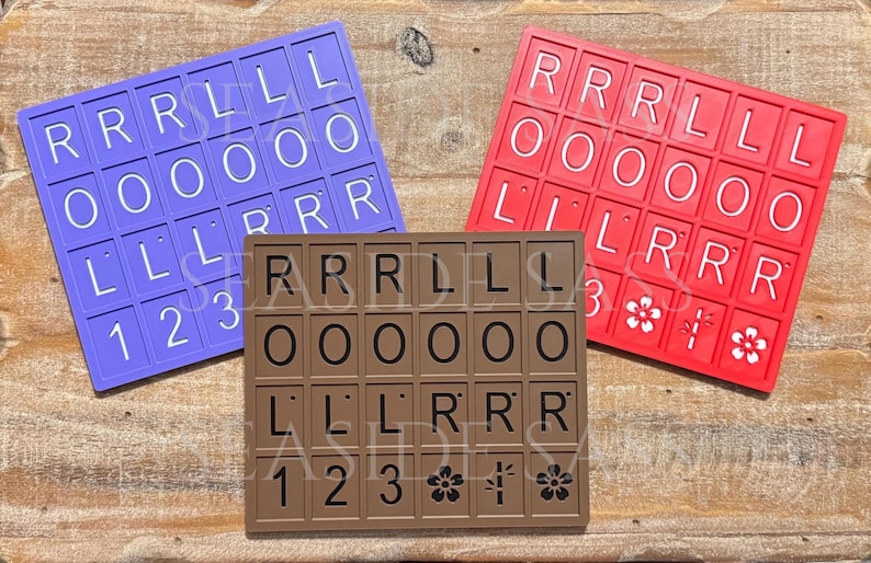 Może przedstawiać: Trzy kolorowe uchwyty na kafelki Scrabble w kolorze fioletowym, czerwonym i brązowym. Każdy uchwyt ma siatkę kwadrat&oacute;w z literami i cyframi. Czerwony uchwyt ma kwiatowe wzory. Litery i cyfry są białe na fioletowych i czerwonych uchwytach i czarne na brązowym uchwycie.