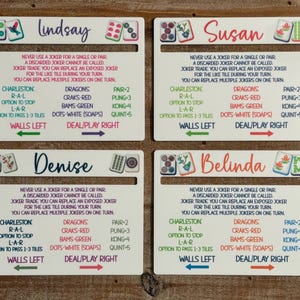 May include: Four white rectangular Mah Jongg card holders with slots, each personalized with names Lindsay, Susan, Denise, and Belinda. The cards feature printed game rules, tile illustrations, and directional arrows for walls and play.