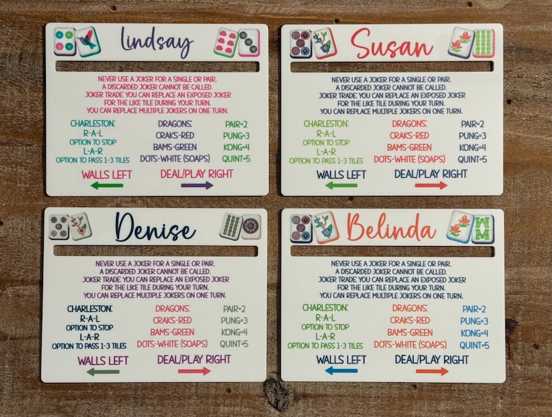May include: Four white rectangular Mah Jongg card holders with slots, each personalized with names Lindsay, Susan, Denise, and Belinda. The cards feature printed game rules, tile illustrations, and directional arrows for walls and play.