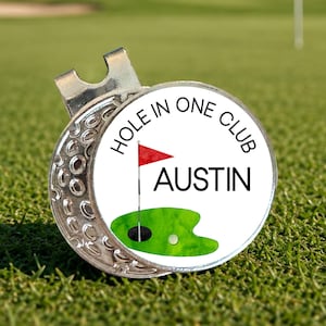 May include: A silver golf ball marker with a circular design. The design features a red flag, a green putting green, and the words "HOLE IN ONE CLUB AUSTIN". The marker is on a green golf course.