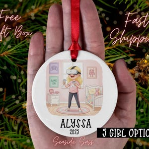 May include: A white ceramic Christmas ornament with a red ribbon. The ornament features a cartoon girl wearing a VR headset, with the name "ALYSSA" and the year "2025" below. The text "5 GIRL OPTIONS" is also visible.