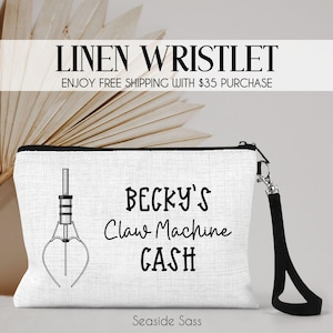May include: White linen wristlet with a black zipper and wrist strap. The wristlet features a claw machine graphic and the text "BECKY'S Claw Machine CASH". The text "LINEN WRISTLET" is at the top.