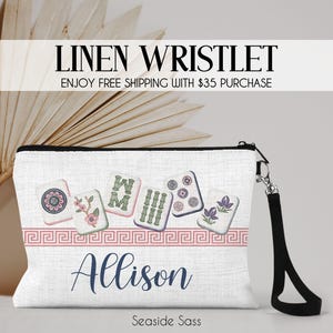 K&ouml;nnte beinhalten: Eine wei&szlig;e Leinen-Handgelenktasche mit einer Reihe von Mahjong-Steinen mit Blumen- und Bambusmotiven, einer rosa M&auml;anderbord&uuml;re und dem Namen Allison in dunkelblauer Schreibschrift. Die Tasche hat eine schwarze Handschlaufe mit silbernem Metallverschluss.