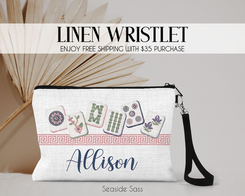 K&ouml;nnte beinhalten: Eine wei&szlig;e Leinen-Handgelenktasche mit einer Reihe von Mahjong-Steinen mit Blumen- und Bambusmotiven, einer rosa M&auml;anderbord&uuml;re und dem Namen Allison in dunkelblauer Schreibschrift. Die Tasche hat eine schwarze Handschlaufe mit silbernem Metallverschluss.