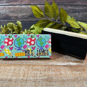 May include: A teacher-themed desk accessory featuring a light blue background with red apple and globe graphics. The name "MRS. JAMES" is printed in black. A black eraser is next to the desk accessory.