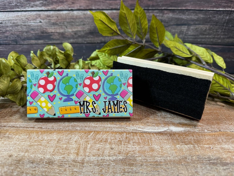 May include: A teacher-themed desk accessory featuring a light blue background with red apple and globe graphics. The name "MRS. JAMES" is printed in black. A black eraser is next to the desk accessory.
