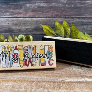 May include: A wooden teacher's stamp with the name "Miss Binford" in cursive, surrounded by colorful school supplies. Next to it is a black chalkboard eraser with a wooden handle. The background is a wooden surface.