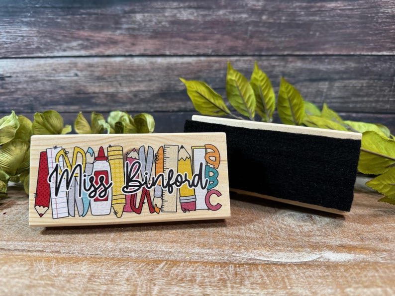 May include: A wooden teacher's stamp with the name "Miss Binford" in cursive, surrounded by colorful school supplies. Next to it is a black chalkboard eraser with a wooden handle. The background is a wooden surface.