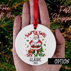 May include: A white Christmas ornament with a red ribbon, featuring Santa Claus and the text "SANTA SAYS SIX SEVEN CLAIRE -2023". The ornament has a festive design with red, green, and brown accents, perfect for holiday decor.