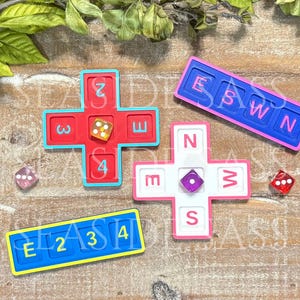 May include: A set of game components including two cross-shaped boards in red and white, two rectangular boards in blue, and three translucent dice. The red board features numbers 2, 3, 4, and E. The white board displays N, E, W, and S. The blue boards show E, 2, 3, 4 and E, S, W, N.