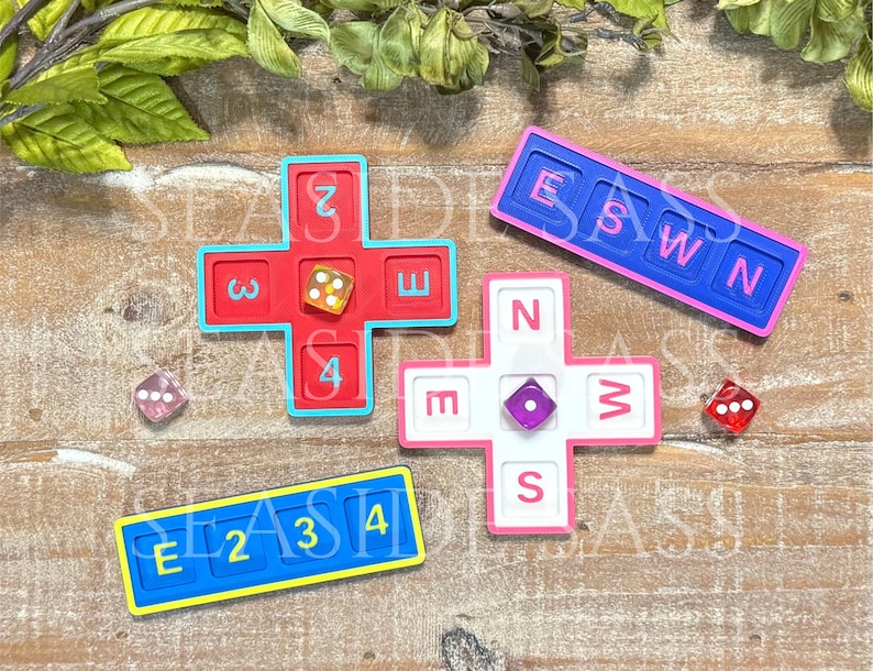 May include: A set of game components including two cross-shaped boards in red and white, two rectangular boards in blue, and three translucent dice. The red board features numbers 2, 3, 4, and E. The white board displays N, E, W, and S. The blue boards show E, 2, 3, 4 and E, S, W, N.