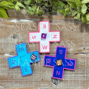 May include: Three cross-shaped game boards in white, light blue, and dark blue, each featuring recessed slots labeled with the letters N, S, E, and W. A single six-sided die rests in the center of each board, displaying different numerical values.