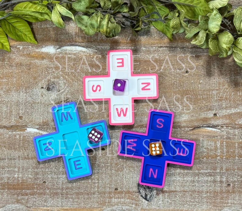 May include: Three cross-shaped game boards in white, light blue, and dark blue, each featuring recessed slots labeled with the letters N, S, E, and W. A single six-sided die rests in the center of each board, displaying different numerical values.