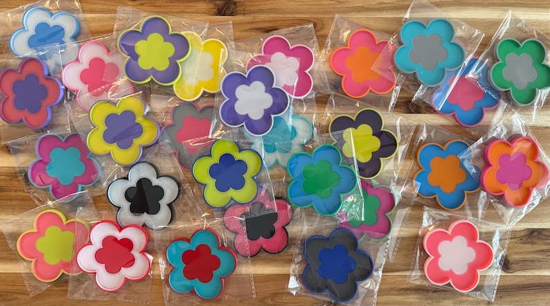 K&ouml;nnte beinhalten: Eine Sammlung bunter, bl&uuml;tenf&ouml;rmiger Untersetzer in verschiedenen Farben, darunter Rosa, Blau, Gelb und Orange. Jeder Untersetzer ist in einer transparenten Plastikt&uuml;te verpackt. Die Untersetzer sind aus strapazierf&auml;higem Material.