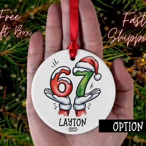 May include: A white ceramic Christmas ornament with the numbers 6 and 7, held by cartoon hands wearing white gloves and red sleeves. The number 7 has a Santa hat. The name "LAYTON" and the year "-2025-" are printed below the numbers. A red ribbon is attached.
