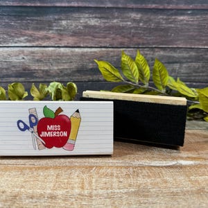 May include: A white rectangular eraser with a black cleaning surface and a wooden handle. Beside it is a white block with school-themed graphics, including an apple, scissors, and a pencil, with the text "MISS JIMERSON".