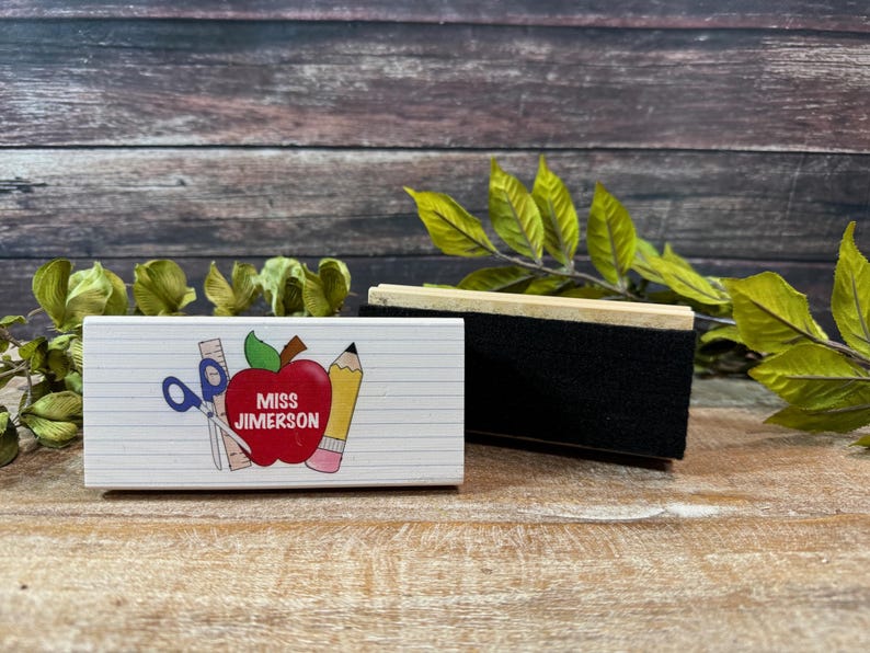 May include: A white rectangular eraser with a black cleaning surface and a wooden handle. Beside it is a white block with school-themed graphics, including an apple, scissors, and a pencil, with the text "MISS JIMERSON".
