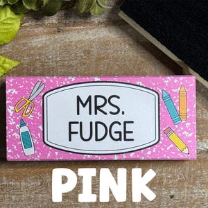 May include: A pink rectangular name tag with the text "MRS. FUDGE" in a white oval. The tag is decorated with school supplies like scissors, glue, crayons, and a pencil. The word "PINK" is written in large white letters at the bottom.