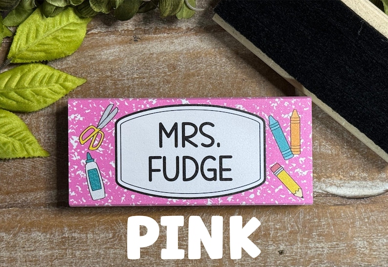 May include: A pink rectangular name tag with the text "MRS. FUDGE" in a white oval. The tag is decorated with school supplies like scissors, glue, crayons, and a pencil. The word "PINK" is written in large white letters at the bottom.