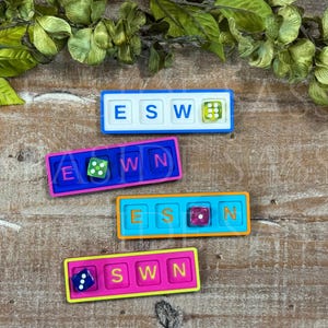 May include: Four rectangular game boards with four square slots each. The boards are white, blue, teal, and pink. Each board contains three letter tiles and one six-sided die in various colors, including green, purple, and blue with white pips.