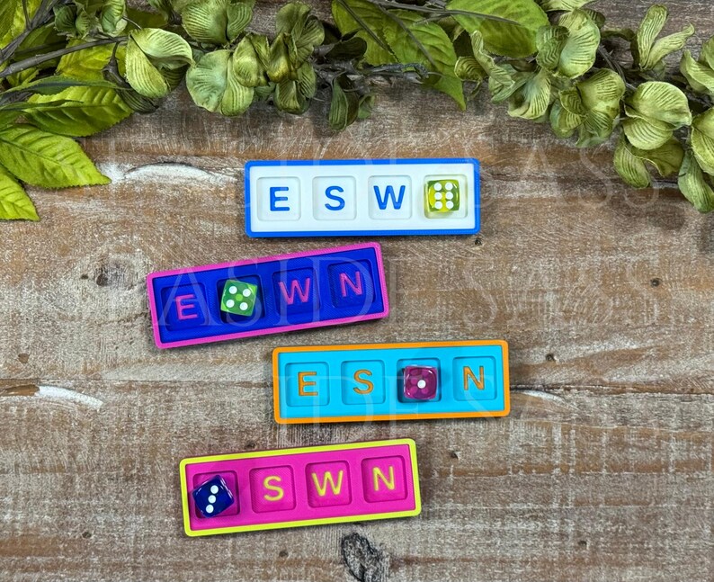 May include: Four rectangular game boards with four square slots each. The boards are white, blue, teal, and pink. Each board contains three letter tiles and one six-sided die in various colors, including green, purple, and blue with white pips.