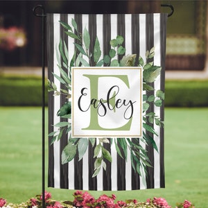 May include: A black and white striped garden flag with green watercolor leaves and a gold frame. The letter "E" is in the center of the frame with the name "Easley" written in cursive below it.