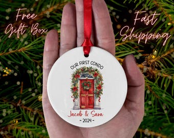 Puede incluir: Un adorno navideño de cerámica blanca con una cinta roja. El adorno presenta una ilustración de acuarela de una puerta roja con una corona floral y el texto "OUR FIRST CONDO" y "Jacob & Sara ~ 2024".