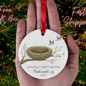 May include: A white ceramic ornament with a red ribbon, featuring a bird's nest on a branch with two birds flying above. The text reads "OFFICIALLY EMPTY NESTERS" with the names "Mark and Liz" and the year "2025".