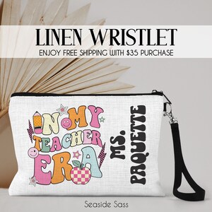 May include: A white linen wristlet with a black zipper and a black strap. The wristlet has a colorful graphic that says "In My Teacher Era" with a pencil, apple, and stars. The text "Ms. Paquette" is printed on the right side of the wristlet.