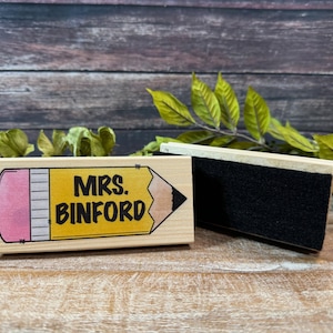 May include: A wooden stamp with a pencil design and the text "MRS. BINFORD" in black. Next to it is a black eraser. The background is a wooden surface with green leaves.