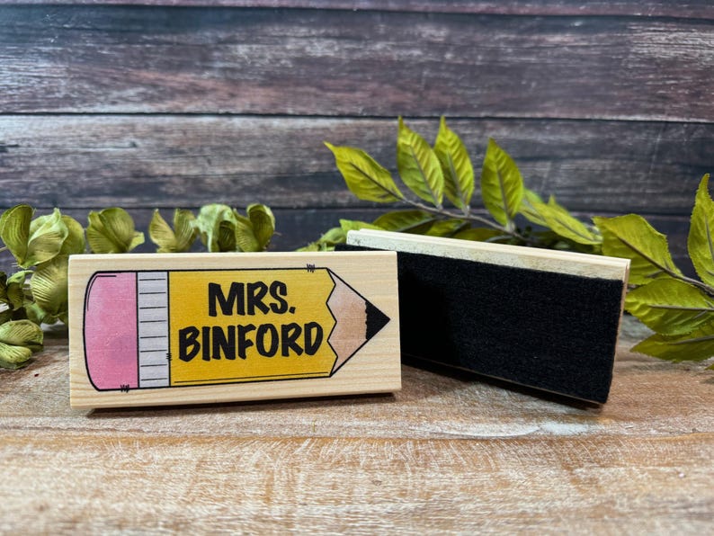 May include: A wooden stamp with a pencil design and the text "MRS. BINFORD" in black. Next to it is a black eraser. The background is a wooden surface with green leaves.
