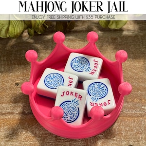 May include: A pink crown-shaped tray holds four white Mahjong tiles with blue circular designs and the word "JOKER" in red. The tray has rounded pink accents. The text "MAHJONG JOKER JAIL" is at the top.