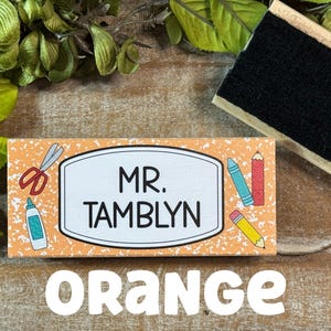 May include: Orange rectangular wooden sign with the name "MR. TAMBLYN" in black. The sign has school-themed illustrations and the word "ORANGE" in large white letters.
