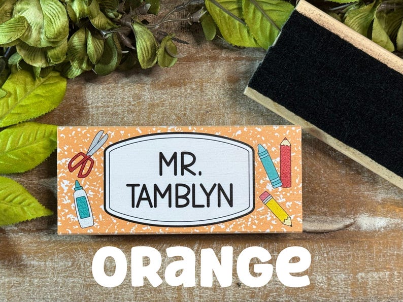 May include: Orange rectangular wooden sign with the name "MR. TAMBLYN" in black. The sign has school-themed illustrations and the word "ORANGE" in large white letters.