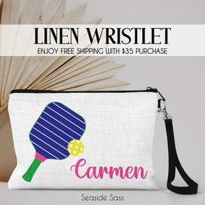May include: White linen wristlet with a black zipper and a black wrist strap. The wristlet features a blue and white striped paddleball with a yellow and white ball and the name "Carmen" in pink lettering. The text "Seashore Sass" is printed in black at the bottom.