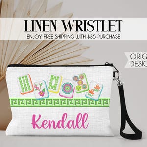K&ouml;nnte beinhalten: Eine wei&szlig;e Leinen-Handgelenktasche mit schwarzer Handschlaufe und einem bunten Grafikdruck von f&uuml;nf Mahjong-Steinen &uuml;ber einer gr&uuml;nen griechischen Bord&uuml;re. Der Name Kendall ist in pinker Schreibschrift auf der Vorderseite aufgedruckt.