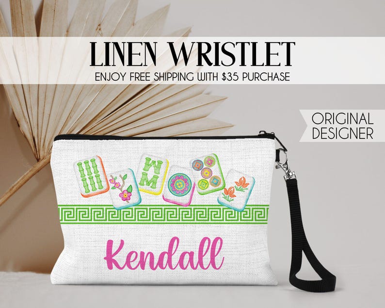 K&ouml;nnte beinhalten: Eine wei&szlig;e Leinen-Handgelenktasche mit schwarzer Handschlaufe und einem bunten Grafikdruck von f&uuml;nf Mahjong-Steinen &uuml;ber einer gr&uuml;nen griechischen Bord&uuml;re. Der Name Kendall ist in pinker Schreibschrift auf der Vorderseite aufgedruckt.