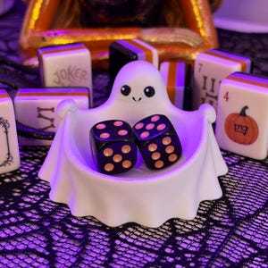 May include: A white ghost-shaped bowl holding two black dice with orange pips, surrounded by several mahjong tiles featuring Halloween-themed designs including a pumpkin, a joker, and ornate frames with orange, black, and white edges.