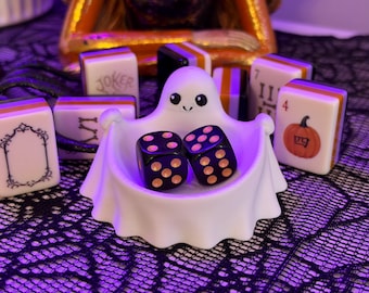 Può includere: Una ciotola bianca a forma di fantasma contenente due dadi neri con puntini arancioni, circondata da diverse tessere del mahjong con disegni a tema Halloween, tra cui una zucca, un jolly e cornici ornate con bordi arancioni, neri e bianchi.