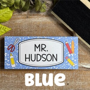 May include: A blue name tag with the text "MR. HUDSON" in black, surrounded by school-themed illustrations. The word "BLUE" is written in white block letters at the bottom. Scissors, glue, and pencils are depicted on the tag.
