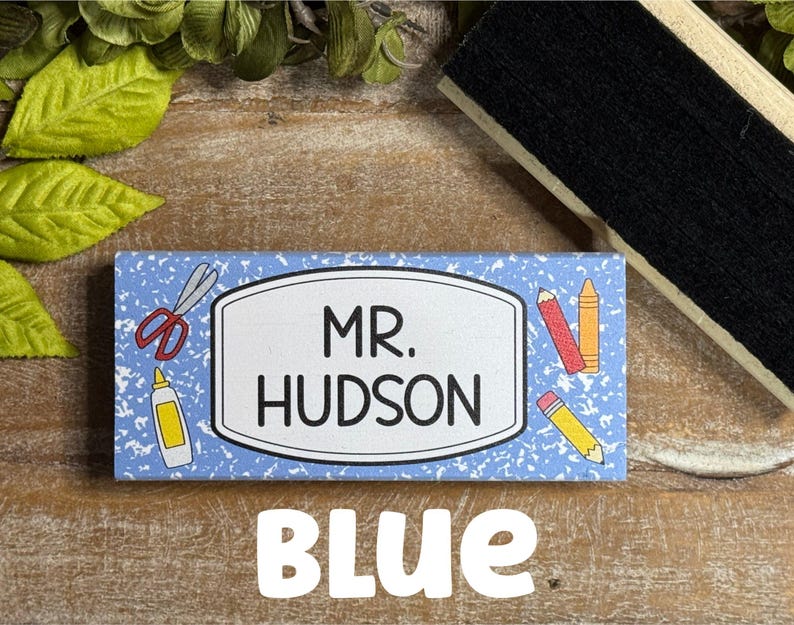 May include: A blue name tag with the text "MR. HUDSON" in black, surrounded by school-themed illustrations. The word "BLUE" is written in white block letters at the bottom. Scissors, glue, and pencils are depicted on the tag.