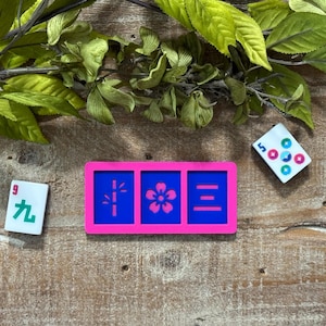 May include: A rectangular pink frame containing three blue panels with pink symbols: a bamboo stalk, a five-petaled flower, and three horizontal lines. Two white mahjong tiles are placed nearby, one with the number 9 and green character, the other with a 5 and colorful circles.