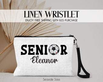 May include: White linen wristlet with a black zipper and a black strap. The wristlet has a black and white soccer ball graphic and the text "Senior Eleanor".