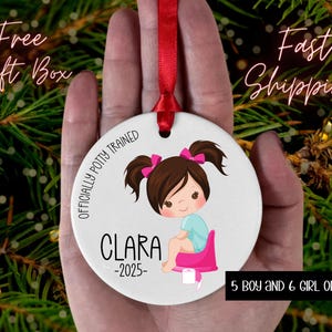 May include: A white ceramic ornament with a red ribbon, featuring a cartoon girl on a potty. The ornament has the text "OFFICIALLY POTTY TRAINED" and "CLARA -2025". Additional text includes "5 BOY AND 6 GIRL OPTIONS".