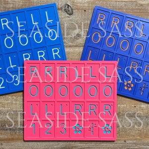 Może przedstawiać: Trzy kolorowe uchwyty na kafelki Scrabble w kolorze niebieskim i r&oacute;żowym. Niebieskie uchwyty mają białe litery i cyfry, a r&oacute;żowy uchwyt ma niebieskie litery i cyfry. Tekst "SEASIDE SASS" jest widoczny na każdym uchwycie.