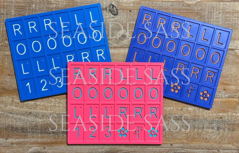 Może przedstawiać: Trzy kolorowe uchwyty na kafelki Scrabble w kolorze niebieskim i r&oacute;żowym. Niebieskie uchwyty mają białe litery i cyfry, a r&oacute;żowy uchwyt ma niebieskie litery i cyfry. Tekst "SEASIDE SASS" jest widoczny na każdym uchwycie.